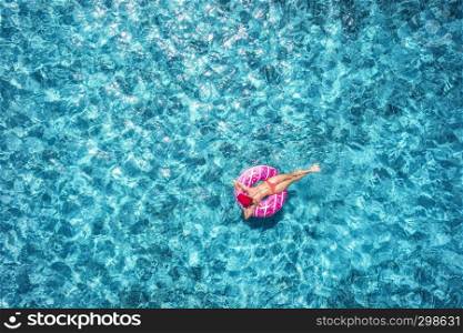Aerial view of slim young woman swimming on the pink donut swim ring in clear blue sea at sunny bright day in summer. Tropical aerial landscape with girl, azure water. Top view from drone. Journey