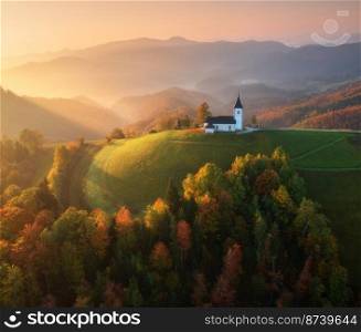 Aerial view of small church in fog and colorful forest at sunrise in autumn. Slovenia. Top view of beautiful chapel on mountain in mist, green meadows, trees, orange sky at dawn in fall. Landscape 