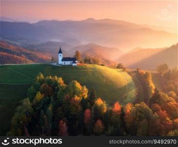 Aerial view of small church in fog and colorful forest at sunrise in autumn. Slovenia. Top drone view of beautiful chapel on mountain in mist, green meadows, trees, pink sky at dawn in fall. Scenery