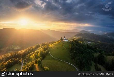 Aerial view of small church on the mountain peak at sunset in∑mer in Slovenia. Top view of beautiful chapel, trail around the hill, green meadows, trees, oran≥sky at twilight in spring. Nature