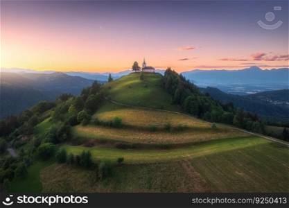 Aerial view of small church on the mountain peak at sunset in summer in Slovenia. Top view of beautiful chapel, trail around the hill, green meadows, trees, orange sky at twilight in spring. Nature