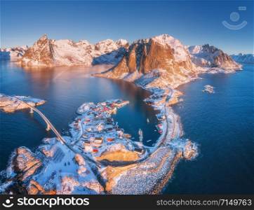 Aerial view of small islands and bridge over the sea and snowy mountains in Lofoten Islands, Norway. Hamnoy at sunrise in winter. landscape with blue water, rocks, buildings, rorbu and road. Top view