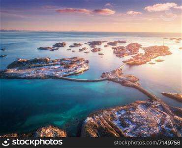 Aerial view of small islands, bridge over the sea and snowy mountains. Lofoten, Norway. Henningsvaer at sunset in winter. landscape with blue water, sky with clouds, rocks, buildings, road. Top view