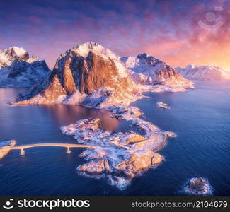 Aerial view of snowy island with rorbu, sea, bridge, rocks in water, mountains, purple sky with pink clouds at sunrise in winter. Beautiful landscape. Top view. Hamnoy village, Lofoten islands, Norway