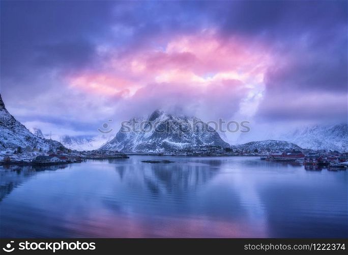 Aerial view of snowy mountain, village on sea coast, purple sky at sunset in winter. Top view of Reine, Lofoten islands, Norway. Moody landscape with high rocks, houses, rorbu, reflection in water