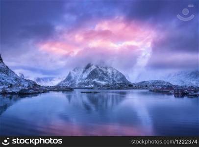Aerial view of snowy mountain, village on sea coast, purple sky at sunset in winter. Top view of Reine, Lofoten islands, Norway. Moody landscape with high rocks, houses, rorbu, reflection in water