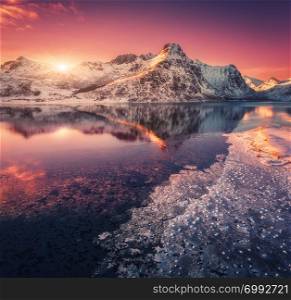 Aerial view of snowy mountains, blue sea with frosty coast, reflection in water and purple sky at colorful sunset in Lofoten islands, Norway. Winter landscape with snow covered rocks, fjord with ice