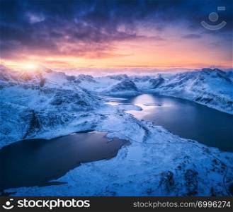 Aerial view of snowy mountains, sea, colorful cloudy sky at sunset in Lofoten islands, Norway. Winter landscape with snow covered rocks and seacoast and blue sky. Top view of Norwegian Fjords at dusk