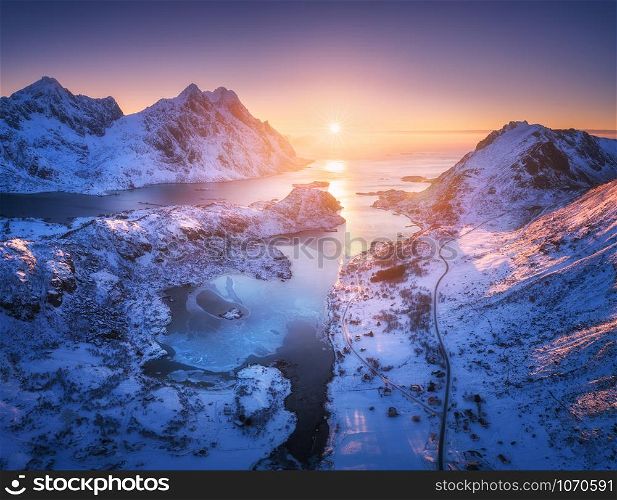 Aerial view of snowy mountains, sea, purple sky at sunset in Lofoten islands, Norway. Winter colorful landscape with snow covered rocks, frosty seacoast, road and village. Top view of Norwegian Fjords