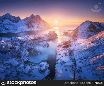 Aerial view of snowy mountains, sea, purple sky at sunset in Lofoten islands, Norway. Winter colorful landscape with snow covered rocks, frosty seacoast, road and village. Top view of Norwegian Fjords
