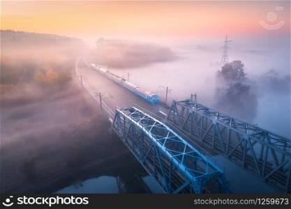 Aerial view of speed train on railroad bridge and river in fog at sunrise in fall. Autumn landscape with foggy fields, mist, water, trees, railway station, orange sky with gold sunlight. Top view