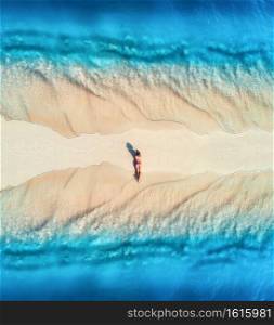 Aerial view of the beautiful young lying woman on the sandy beach and blue sea with waves on the both sides at sunset. Summer holiday. Top view of the back of sporty slim girl, sandbank, clear water