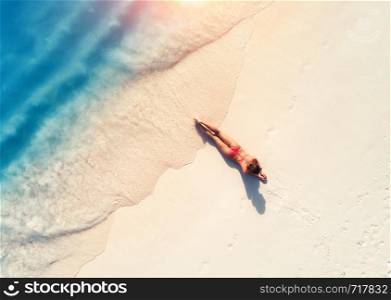 Aerial view of the beautiful young lying woman on the sandy beach near blue sea with waves at sunset. Summer holiday. Top view of the back of sporty slim girl, clear water. Sexy buttocks. Relax. Aerial view of the beautiful young lying woman on the sandy beac