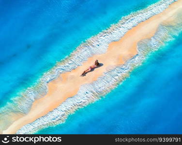 Aerial view of the beautiful young lying woman on the sandy beach and blue sea with waves on the both sides at sunset. Summer holiday. Top view of the back of sporty slim girl, sandbank, clear water