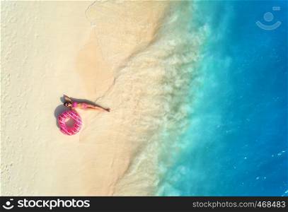 Aerial view of the beautiful young lying woman with pink donut swim ring on the white sandy beach near blue sea with waves at sunset. Summer holiday. Top view of slim girl, clear water. Indian Ocean