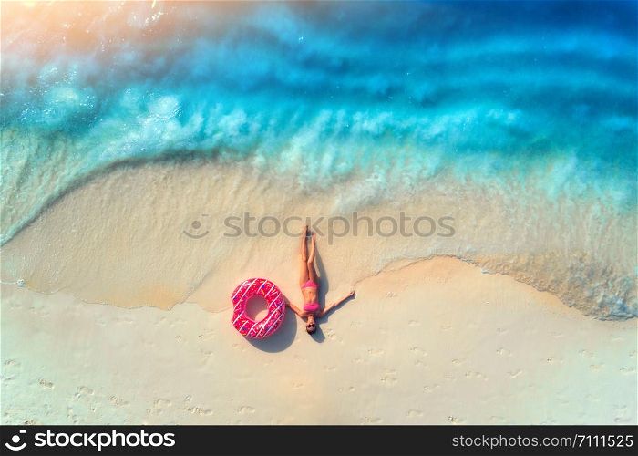 Aerial view of the beautiful young lying woman with pink donut swim ring on the white sandy beach near sea with waves at sunset. Summer holiday. Top view of slim girl, clear azure water. Indian Ocean