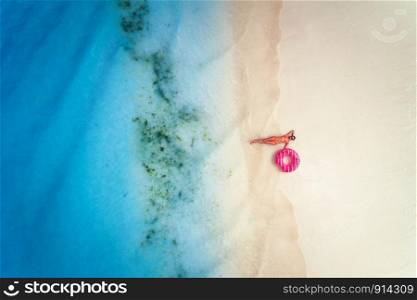 Aerial view of the beautiful young lying woman with pink donut swim ring on white sandy beach near blue sea with waves at sunset. Tropical summer holiday. Top view of slim girl, clear water. Leisure