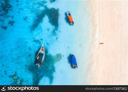 Aerial view of the fishing boats in clear blue water at sunset in summer. Top view from of boat, yacht, sea and walking man on the sandy beach in Zanzibar. Travel. Tropical seascape. View from above. Aerial view of the fishing boat in clear blue water at sunset