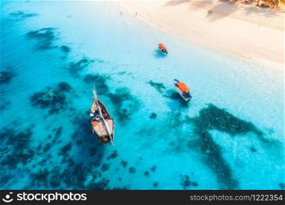 Aerial view of the fishing boats in transparent blue water at sunset in summer. Top view from drone of floating boat in Indian ocean in Zanzibar, Africa. Landscape with yachts in clear sea. Seascape