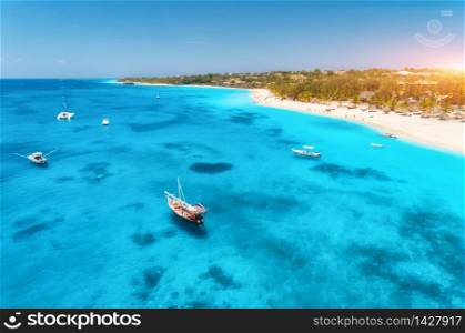 Aerial view of the fishing boats on tropical sea coast with sandy beach at sunrise. Summer holiday in Zanzibar, Africa. Landscape with boat, yacht, clear blue water, green palm trees. View from above. Aerial view of the fishing boats on the tropical sea coast