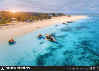 Aerial view of the fishing boats on tropical sea coast with sandy beach at sunset. Summer holiday in Zanzibar, Africa. Landscape with boat, yacht in clear blue water, green palm trees. View from above