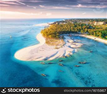 Aerial view of the fishing boats on tropical sea coast with sandy beach at sunset. Summer travel in Zanzibar, Africa. Top view of boats, yachts, green palm trees, clear blue water, colorful sky