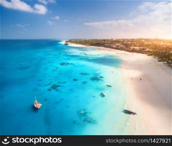 Aerial view of the fishing boats on tropical sea coast with white sandy beach at sunset. Summer travel in Zanzibar, Africa. Landscape with boat, yacht, clear blue water, green palm trees. Top view. Aerial view of the fishing boats on the tropical sea coast