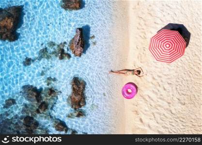 Aerial view of the lying beautiful woman with pink swim ring on the sandy beach, blue sea, stones, red umbrella at sunset. Summer vacation. Lefkada island, Greece. Top view of slim girl, azure water