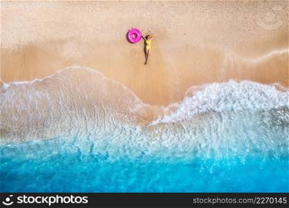Aerial view of the lying beautiful young woman with pink swim ring on the sandy beach near sea with waves at sunset. Summer vacation in Lefkada island, Greece. Top view of slim girl, clear azure water