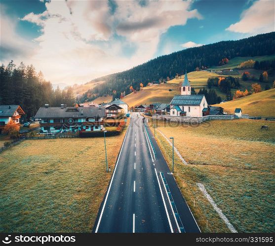 Aerial view of the road in beautiful village in mountain valley at sunset. Dolomites, Italy. Top view of asphalt roadway, church, buildings, meadows grass, trees in autumn. Aerial rural landscape