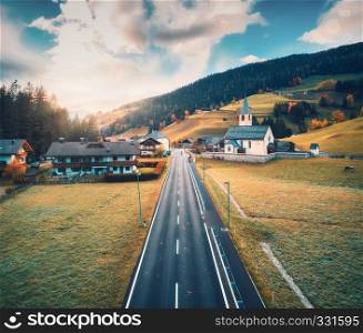 Aerial view of the road in beautiful village in mountain valley at sunset. Dolomites, Italy. Top view of asphalt roadway, church, buildings, meadows grass, trees in autumn. Aerial rural landscape