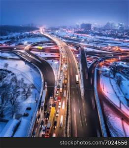 Aerial view of the road in modern city at night in fog. Top view of traffic in highway. Winter cityscape with elevated road, cars, buildings, illumination. Interchange overpass in Europe. Expressway