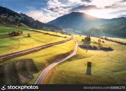 Aerial view of the road in mountain valley at sunrise in Dolomites, Italy. Top view of asphalt roadway, meadows with green grass and trees, buildings, hills in autumn. Highway through the fields