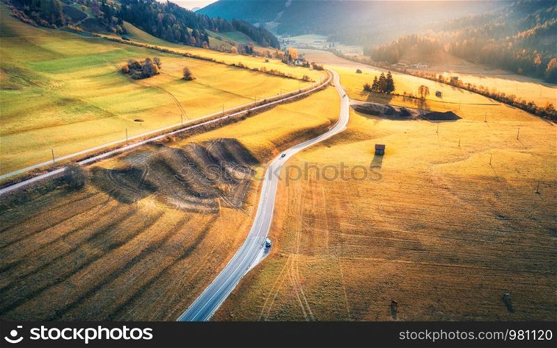 Aerial view of the road in mountain valley at sunset in autumn. Top view of asphalt roadway, railroad, hills with orange meadows, sunlight, yellow trees, buildings. Highway and fields in fall. Scenery. Aerial view of the road in mountain valley at sunset in autumn