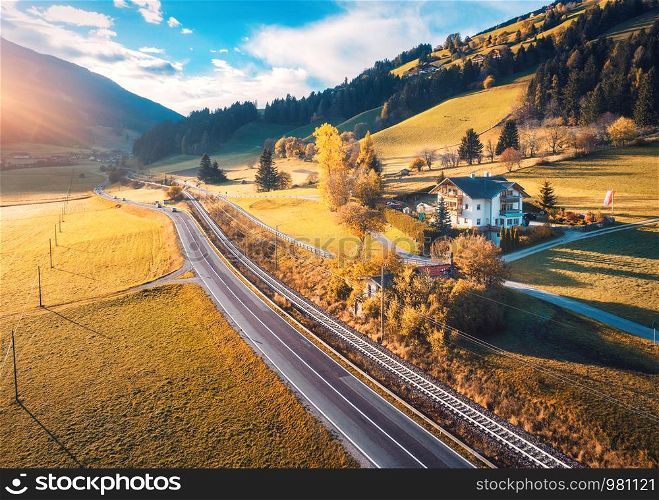 Aerial view of the road in mountain valley at sunset in autumn. Top view of cars on asphalt roadway, house, railroad, hills with orange meadows, blue sky, yellow trees, buildings in fall. Landscape. Aerial view of the road in mountain valley at sunset in autumn