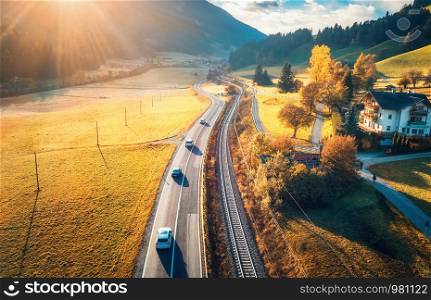 Aerial view of the road in mountain valley at sunset in autumn. Top view of cars on asphalt roadway, house, railroad, hills with orange meadows, blue sky, yellow trees, buildings in fall. Landscape. Aerial view of the road in mountain valley at sunset in autumn
