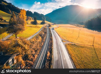 Aerial view of the road in mountain valley at sunset in autumn. Top view of asphalt roadway, railroad, hills with orange meadows, sunlight, yellow trees, buildings. Highway and fields in fall. Scenery. Aerial view of the road in mountain valley at sunset in autumn