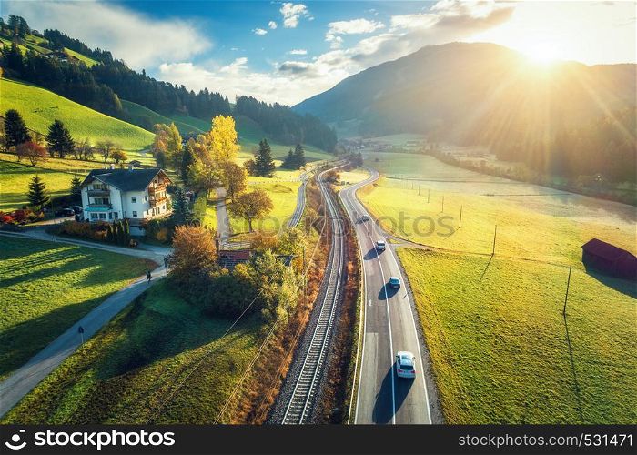 Aerial view of the road in mountain valley at sunset in spring in Dolomites, Italy. Top view of cars on asphalt roadway, house, railroad, hills with green meadows, blue sky, trees, buildings. Highway