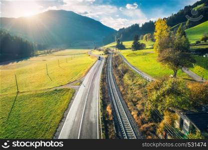 Aerial view of the road in mountain valley at sunset in spring in Dolomites, Italy. Top view of asphalt roadway, railroad, hills with green meadows, blue sky, trees, buildings. Highway and fields