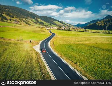 Aerial view of the road in mountain valley at sunset in spring in Dolomites, Italy. Top view of asphalt roadway, car, hills with green meadows, blue sky, trees, buildings. Highway and fields. Nature