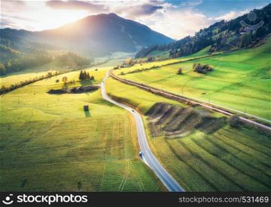 Aerial view of the road in mountain valley at sunset in summer in Dolomites, Italy. Top view of asphalt roadway, railroad, hills with green meadows, blue sky, trees, buildings. Highway and fields