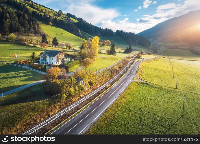 Aerial view of the road in mountain valley at sunset in summer in Dolomites, Italy. Top view of cars on asphalt roadway, house, railroad, hills with green meadows, blue sky, trees, buildings. Highway