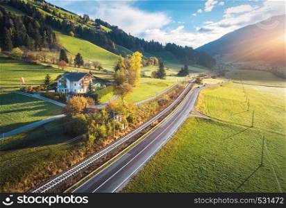 Aerial view of the road in mountain valley at sunset in summer in Dolomites, Italy. Top view of cars on asphalt roadway, house, railroad, hills with green meadows, blue sky, trees, buildings. Highway