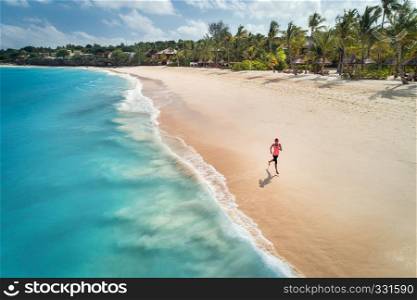 Aerial view of the running young woman on the white sandy beach near sea with waves at sunrise. Summer holiday. Top view of sporty slim girl, clear azure water. Indian Ocean. Lifestyle and sport