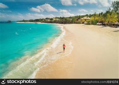 Aerial view of the running young woman on the white sandy beach near sea with waves at sunny day. Summer holiday. Top view of sporty slim girl, clear azure water. Indian Ocean. Lifestyle and sport