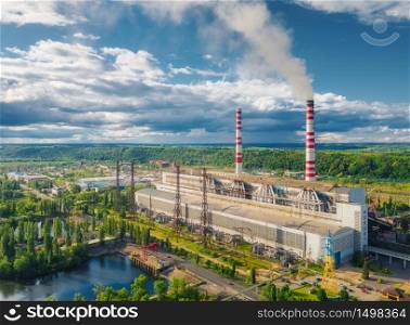 Aerial view of thermal power station. Industrial landscape with thermal power plant, pipes with smoke, buildings, green trees, blue sky with clouds at sunset in summer. Top view. Smoke stack