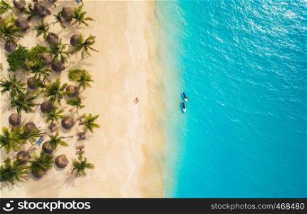 Aerial view of umbrellas, palms on the sandy beach and kayaks in the sea at sunset. Summer holiday in Zanzibar, Africa. Tropical landscape with palm trees, parasols, boat, sand, blue water. Top view
