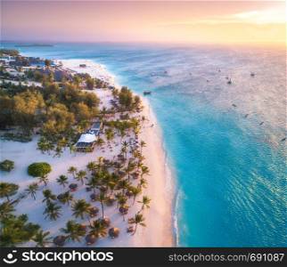 Aerial view of umbrellas, palms on the sandy beach of the sea at sunset. Summer travel in Zanzibar, Africa. Tropical landscape with palm trees, boats, yachts, blue water, orange sky. Top view from air