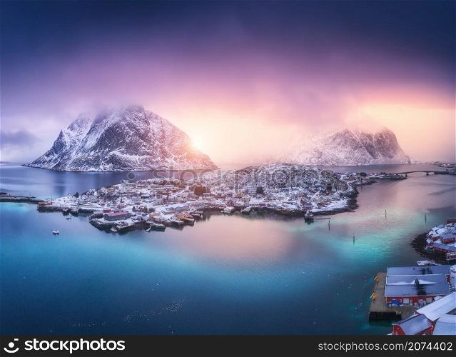 Aerial view of village on the island, blue sea, snowy mountains, sky, violet clouds at colorful sunset in winter. Top view from drone of Reine, Lofoten islands, Norway. Sea coast, city, rocks in fog