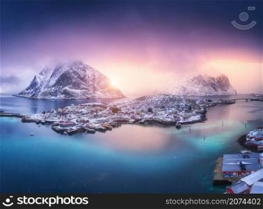 Aerial view of village on the island, blue sea, snowy mountains, sky, violet clouds at colorful sunset in winter. Top view from drone of Reine, Lofoten islands, Norway. Sea coast, city, rocks in fog